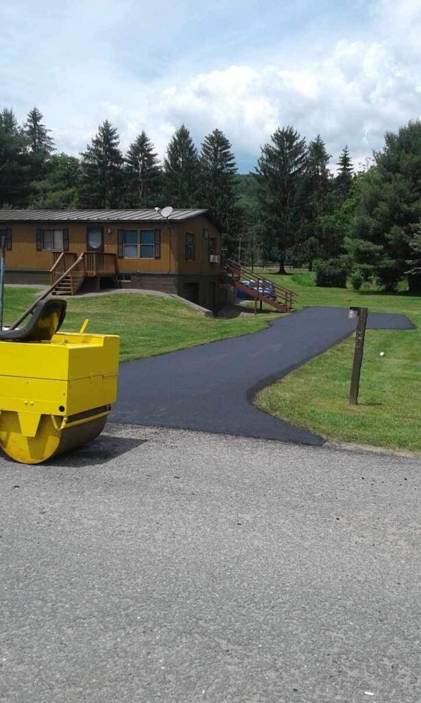 A freshly paved driveway leads to a house surrounded by green lawns and trees. A yellow steamroller is on the road nearby.
