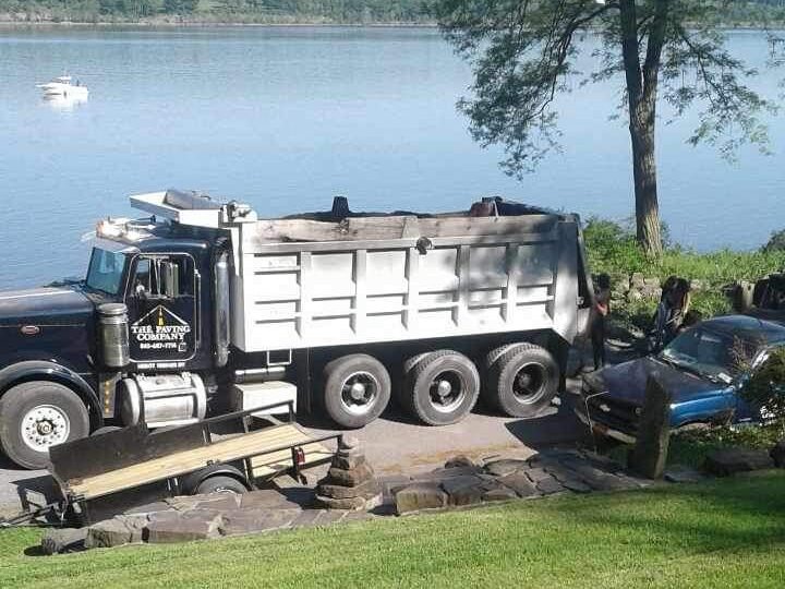 A dump truck labeled "The Paving Company" and a small trailer are parked next to a car near a lake with a tree and stone wall in the background.